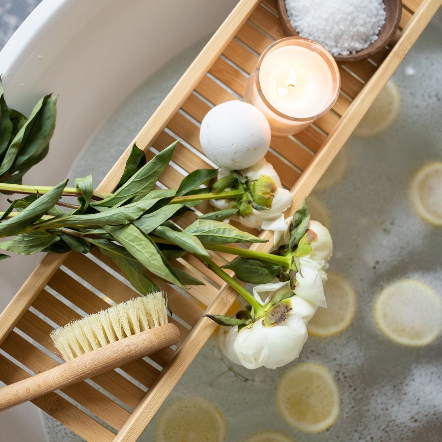 A wooden tray with a bath bomb, salts and a candle over a bathtub with lemons floating in the water
