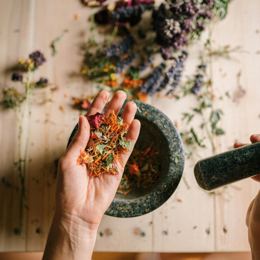 A hand holding herbs and florals over a mortar and pestle 