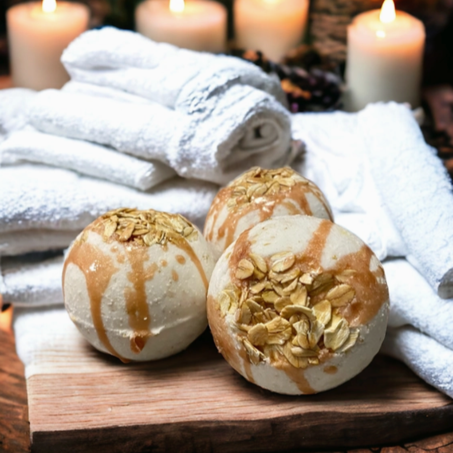 Three oatmeal-inspired bath bombs with golden drizzle and rolled oats beside soft white towels and glowing candles.
