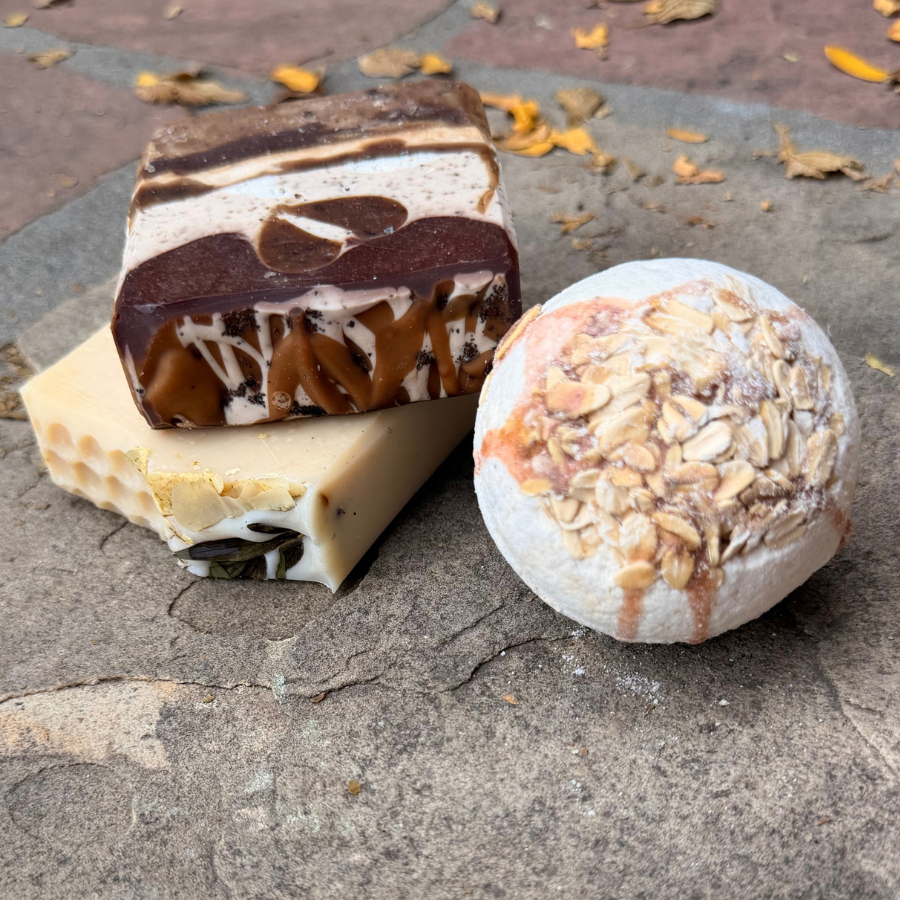 Three artisanal soaps displayed outdoors on a stone surface: a layered chocolate and cream bar, a pale yellow honeycomb-patterned bar, and a round oatmeal-topped soap.
