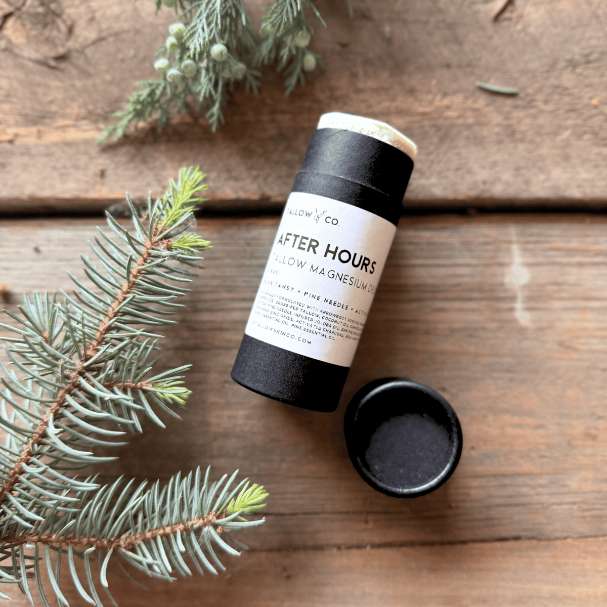 After Hours natural tallow magnesium deodorant with its cap removed, surrounded by pine branches and greenery on a rustic wooden background.