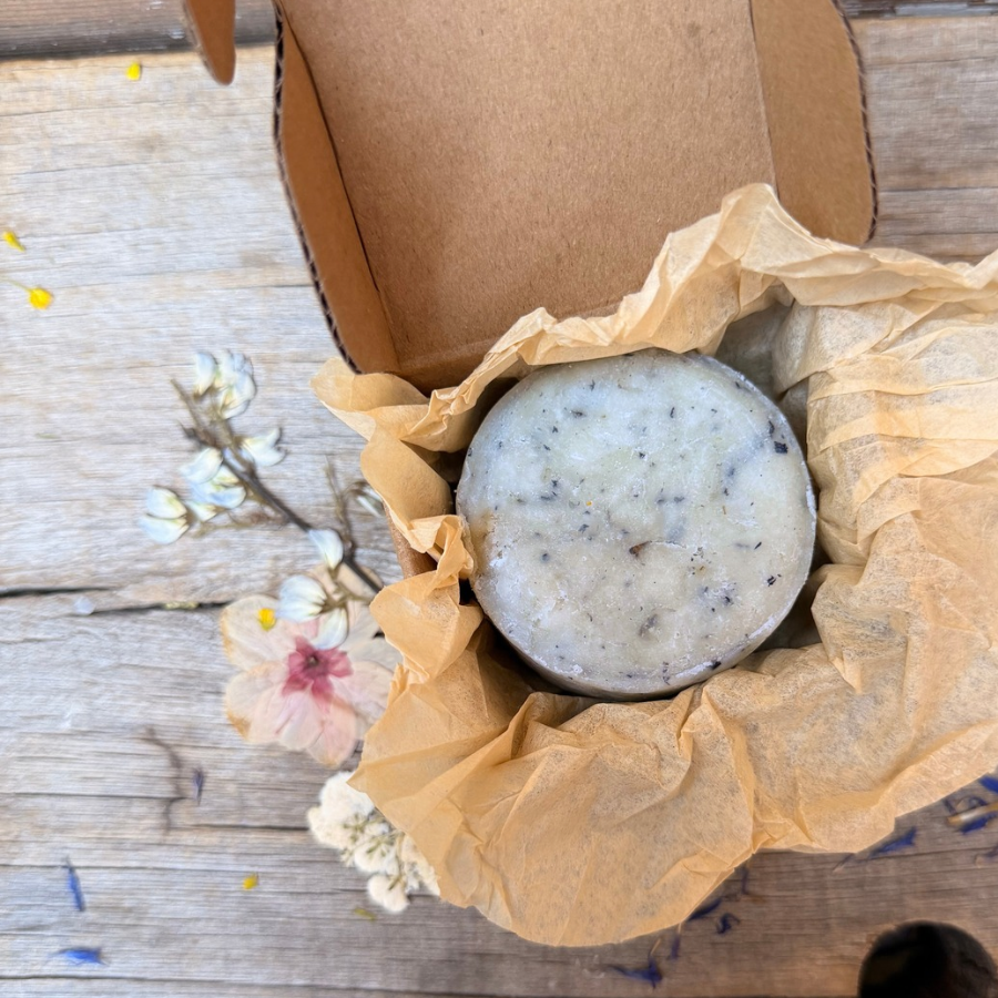 Round Butterfly Pea Facial Cleansing Bar with botanical flecks nestled in kraft tissue inside an open cardboard box.