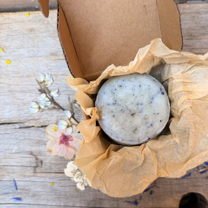 Round Butterfly Pea Facial Cleansing Bar with botanical flecks nestled in kraft tissue inside an open cardboard box.