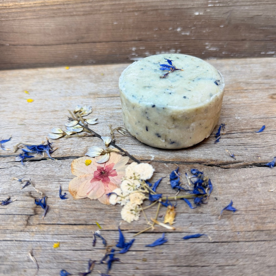 Round Butterfly Pea Facial Cleansing Bar with blue botanical flecks and flower petals, photographed beside dried flowers on a wooden surface.