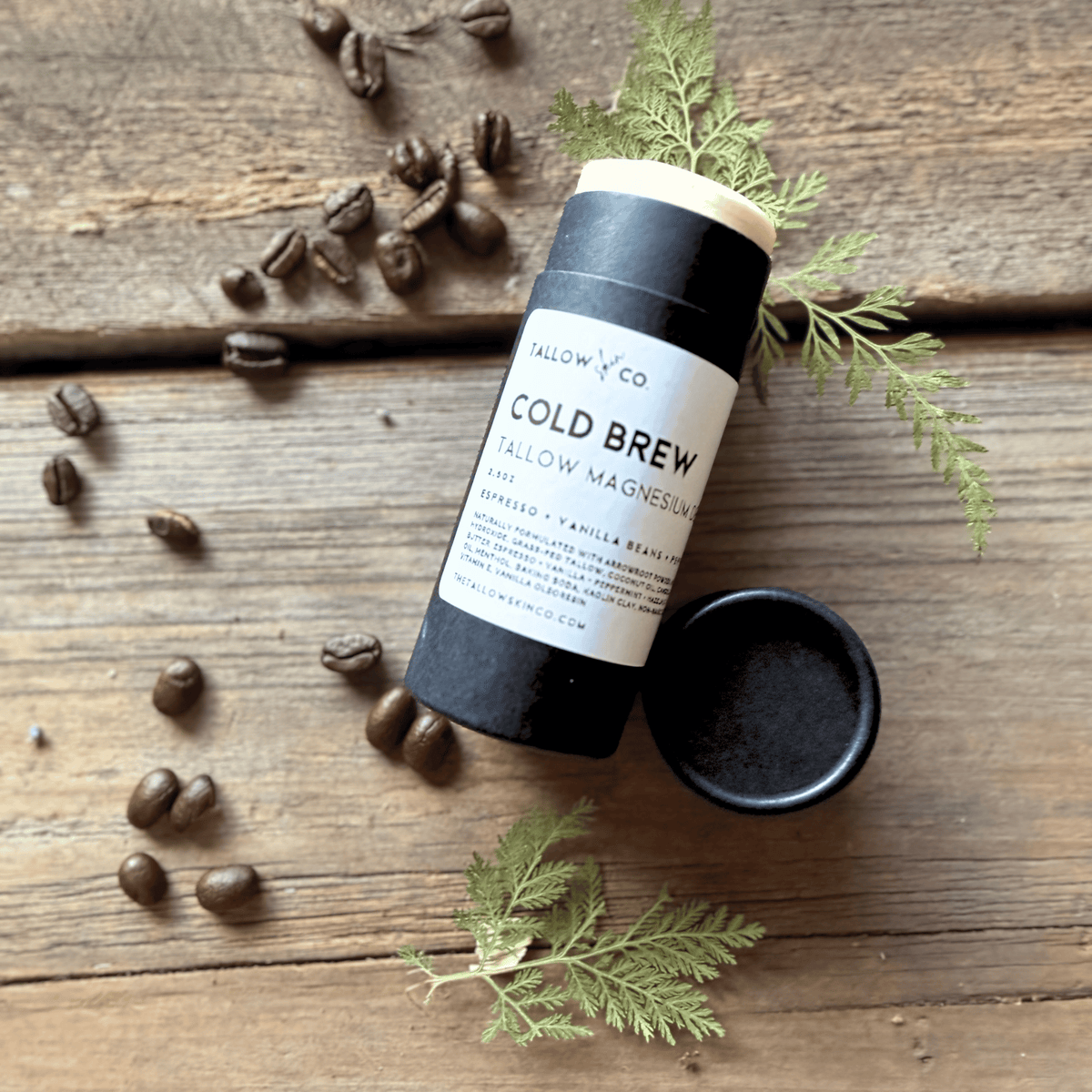 Cold Brew Cooling Tallow Magnesium Deodorant with its cap removed, styled with roasted coffee beans and green botanical sprigs on rustic wooden planks.