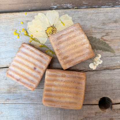 Overhead view of three Wild Blueberry Kombucha Tallow Facial Soap bars with ridged tops, surrounded by pressed flowers on a rustic wooden surface.