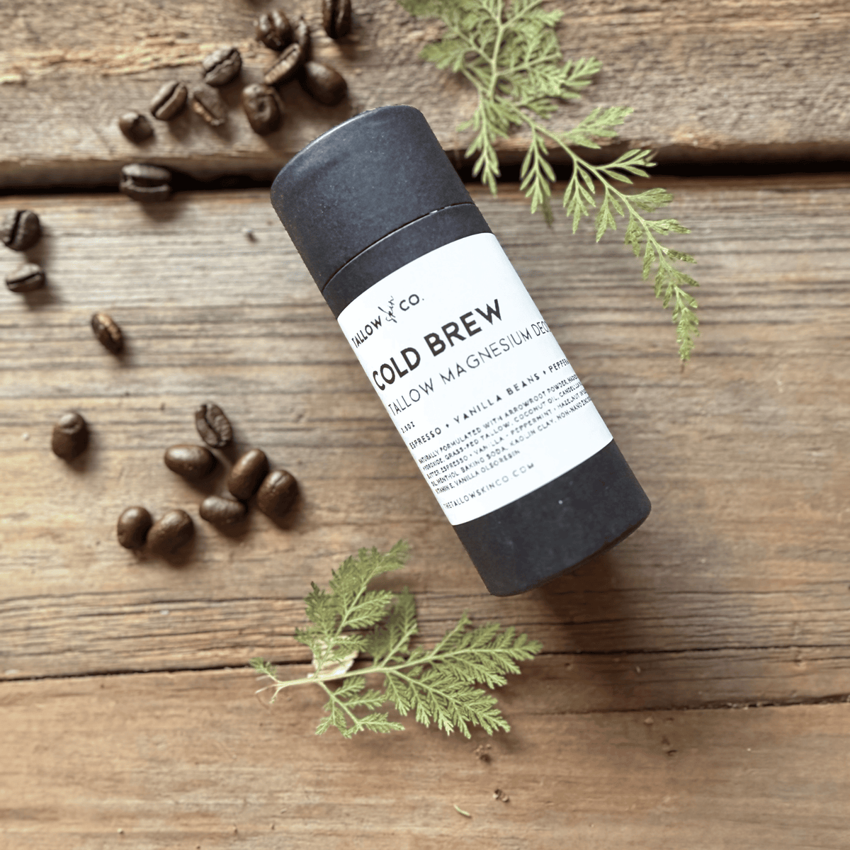 Cold Brew Tallow Magnesium Deodorant in a black paperboard tube, surrounded by roasted coffee beans and fresh greenery on rustic wood.