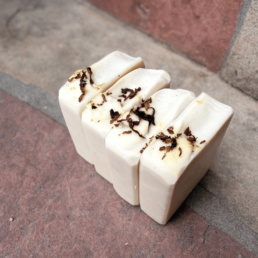 Angled view of four Tobacco & Bay Leaf exfoliating tallow soap bars featuring creamy white tops decorated with real pipe tobacco shavings.